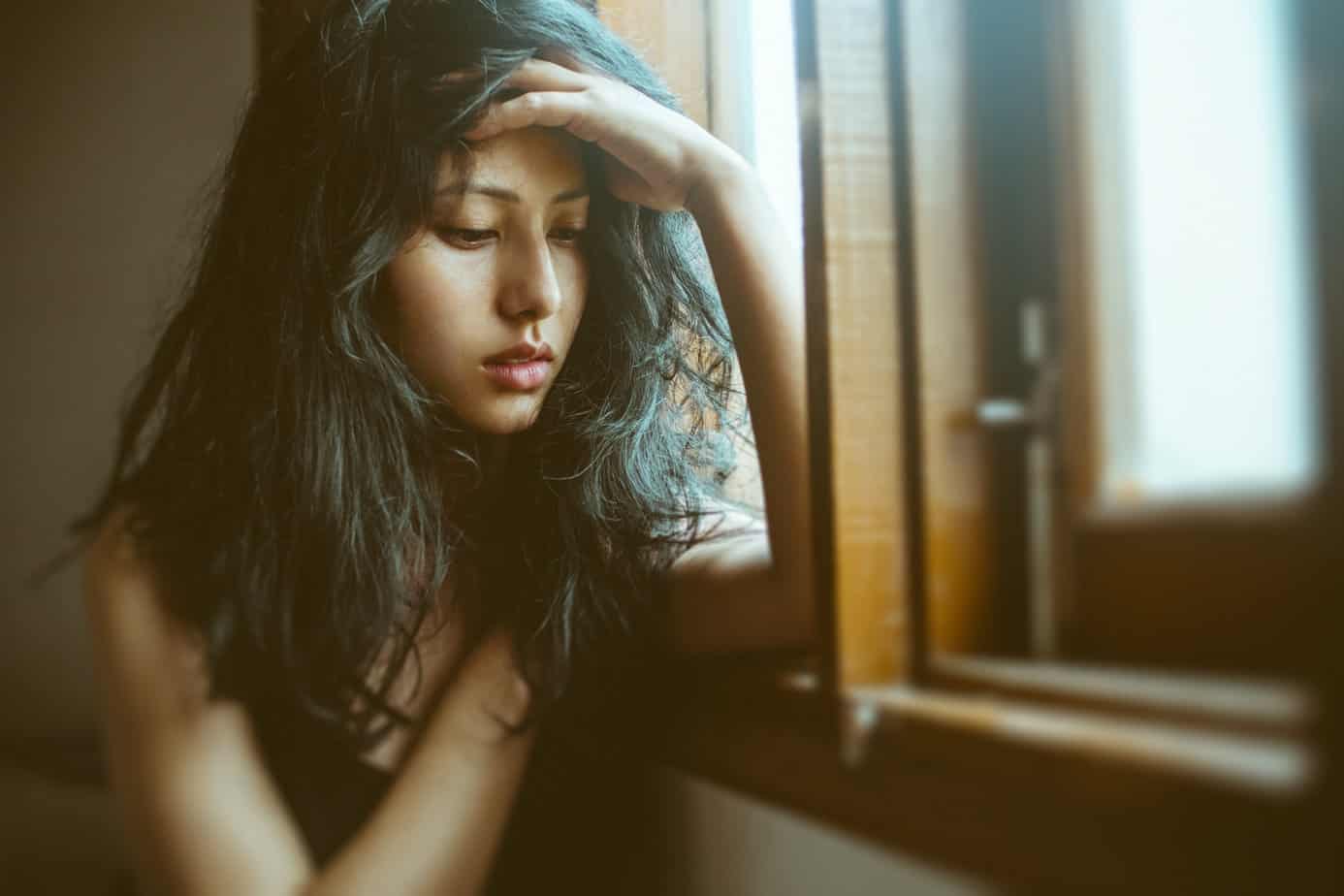 distressed young woman in drug withdrawal sits by window with head in hands