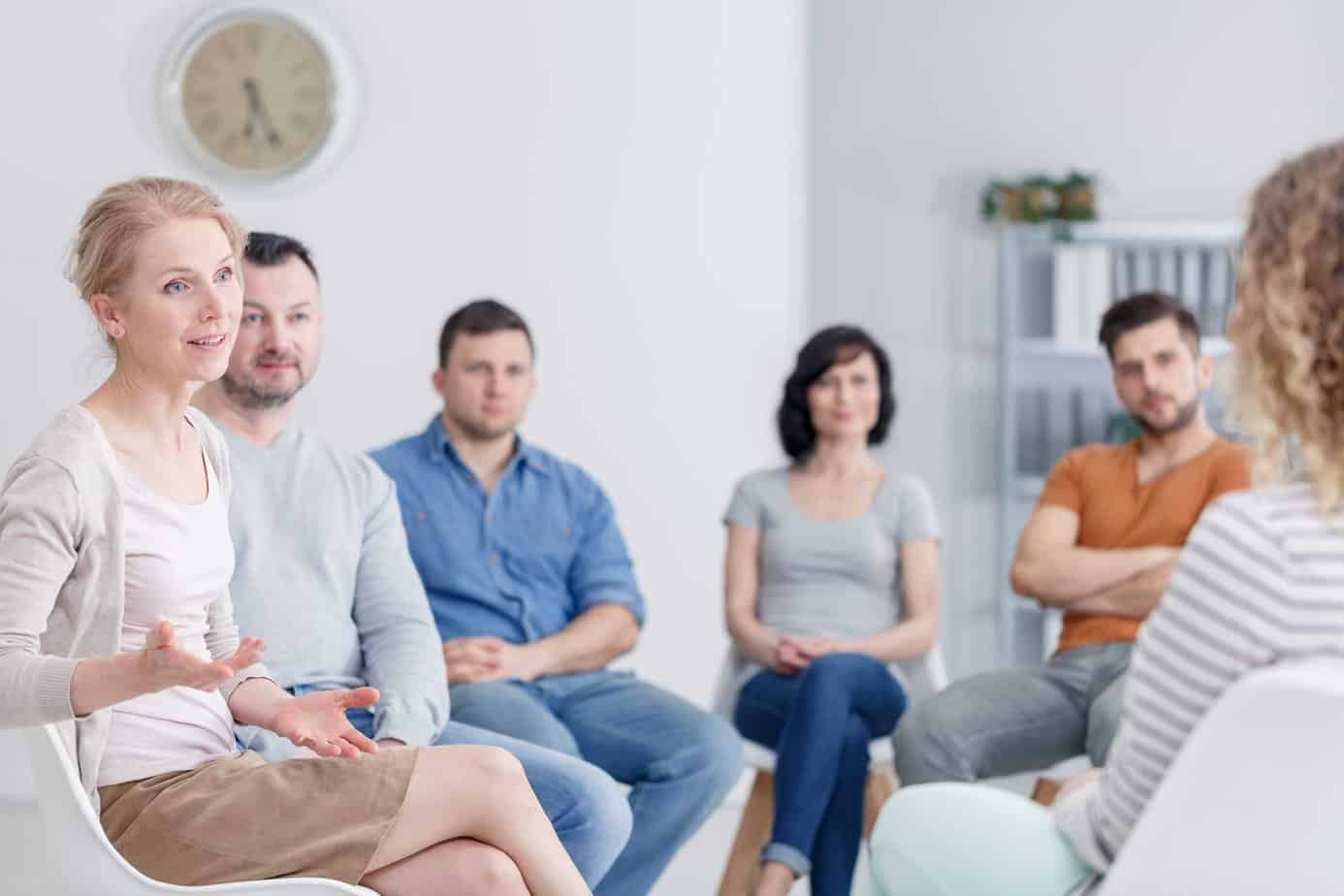 people seated in recovery meeting, with woman talking to others