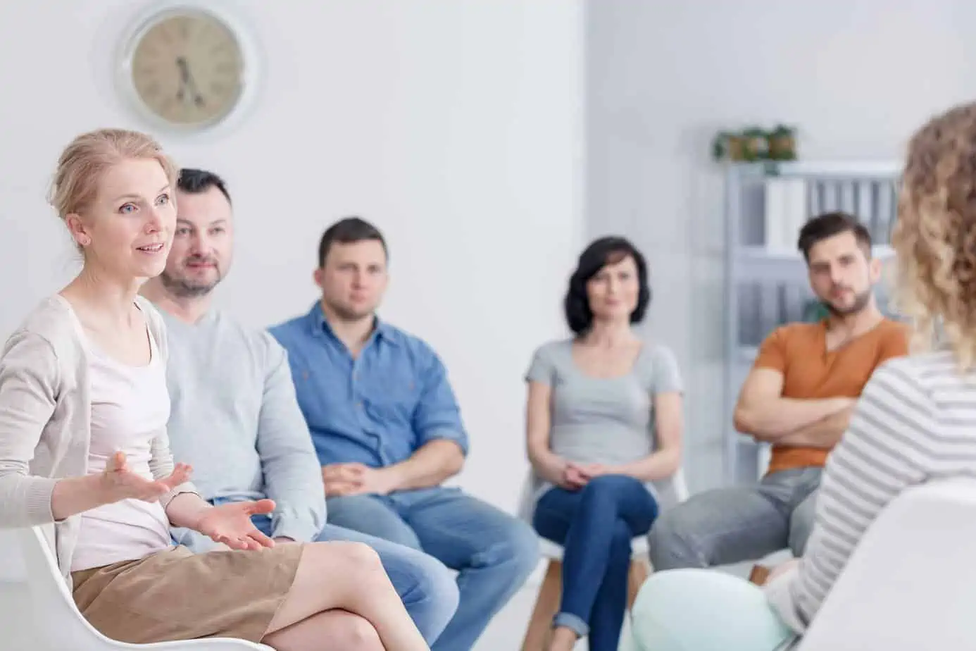 people seated in recovery meeting, with woman talking to others
