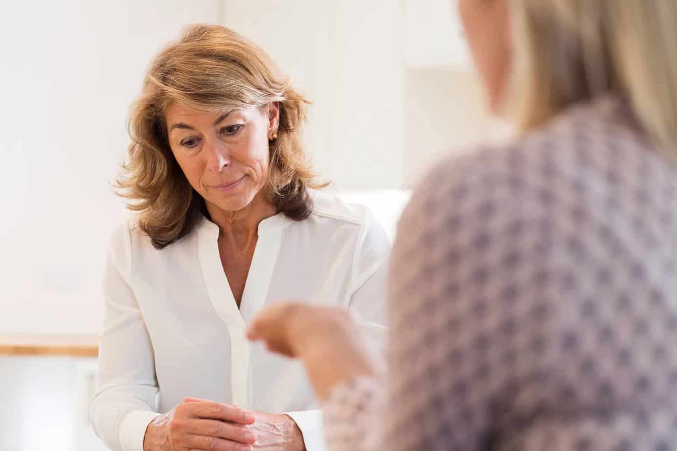 older blonde woman in white shirt looking down hands folded with another woman across from her talking about rehab
