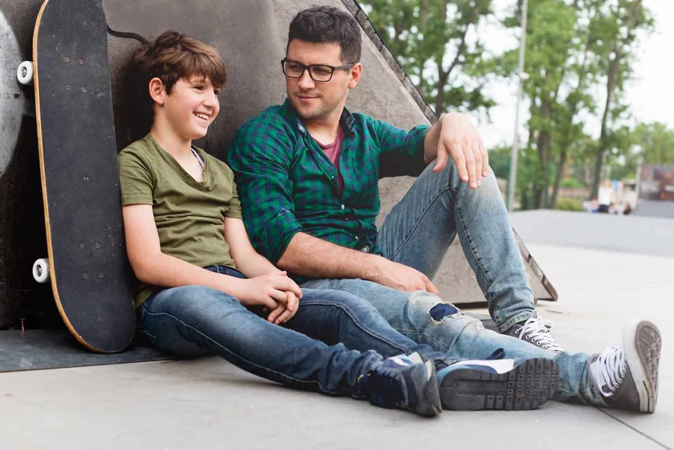 Man and boy sitting next to skateboard in park smiling and talking