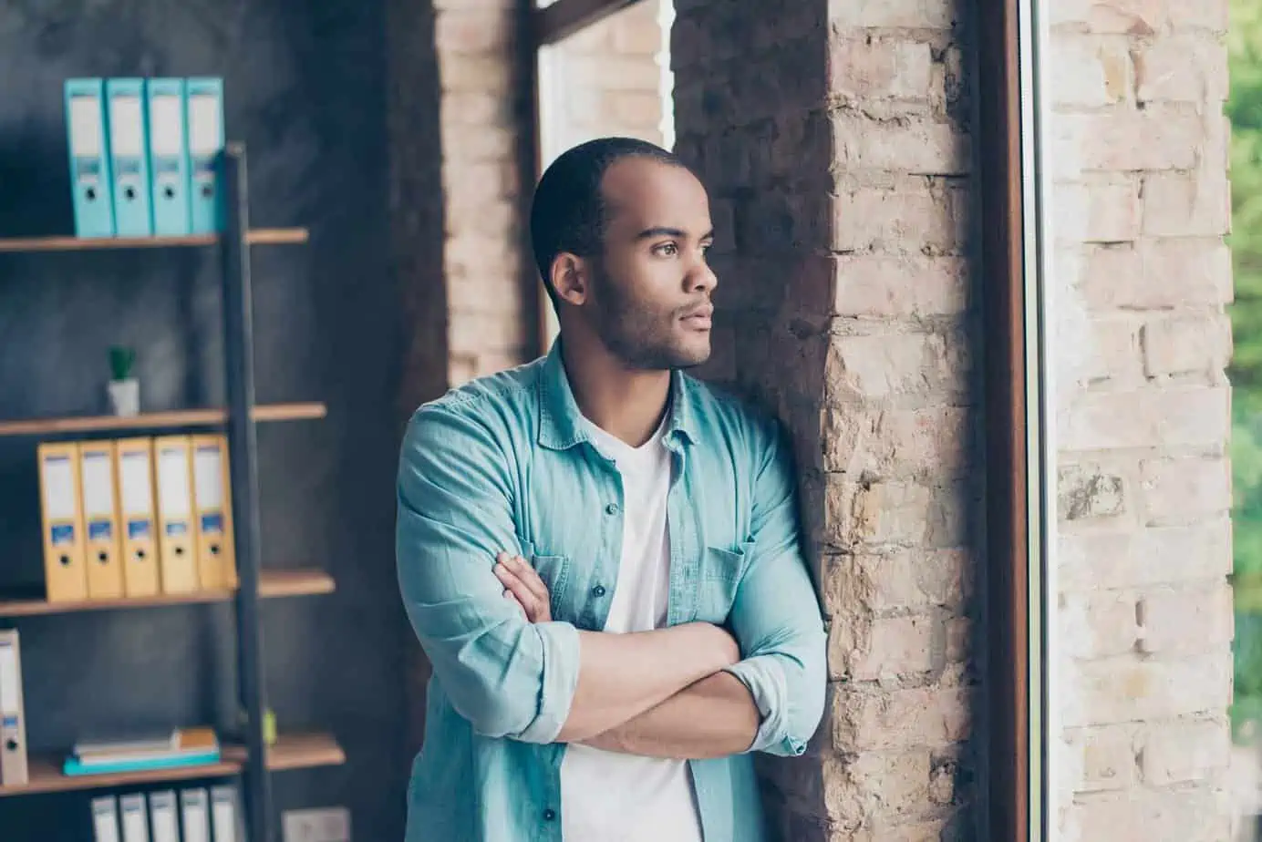 Man leaning on brick wall looking at window with arms crossed in thought