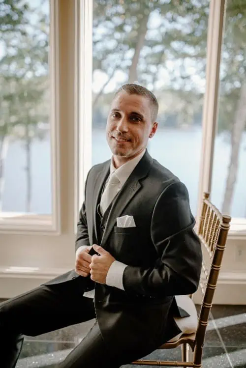 man in tuxedo smiles in chair next to window with lake view